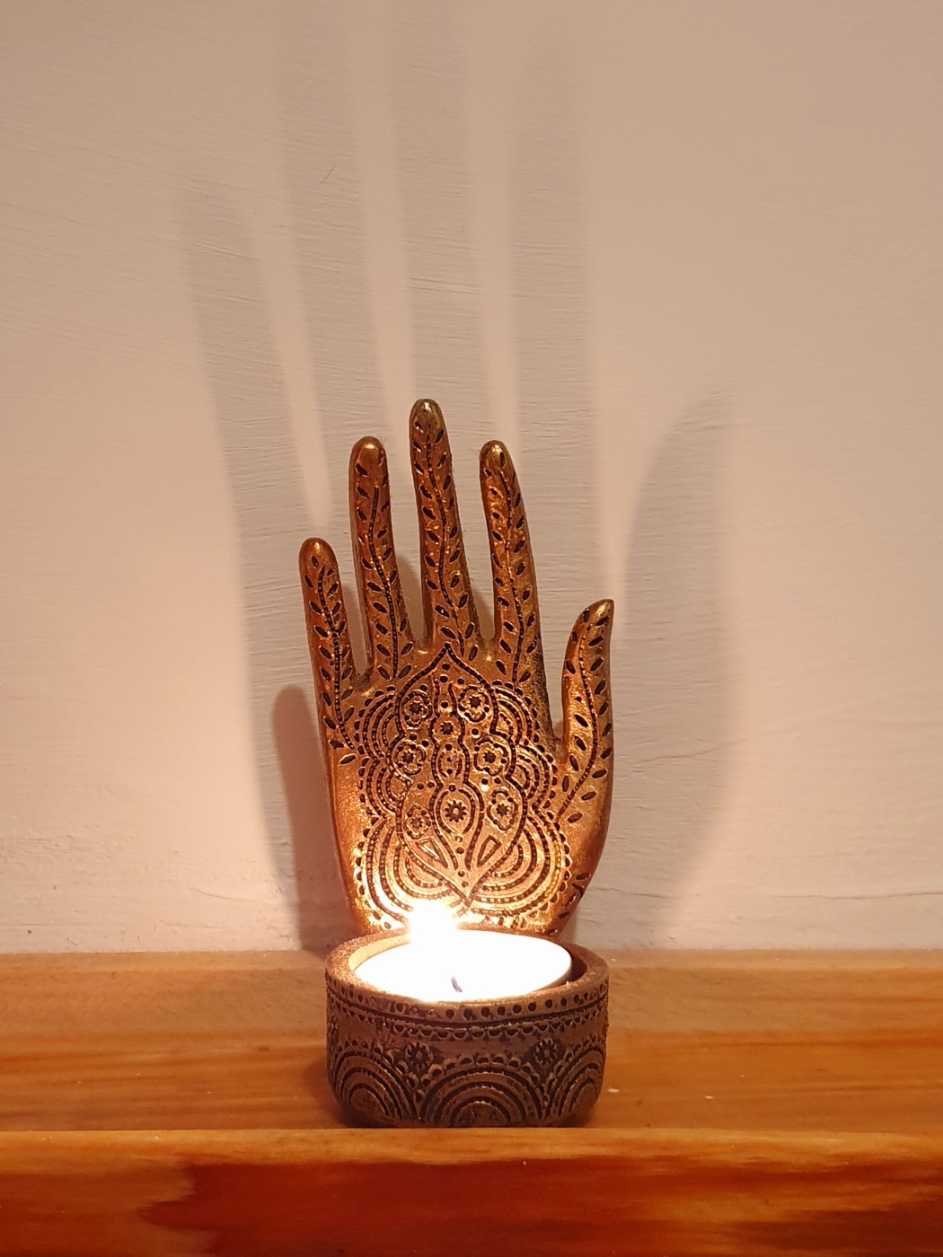 A brass hand-shaped candle holder with a lit tea light, on a wooden shelf against a pale wall