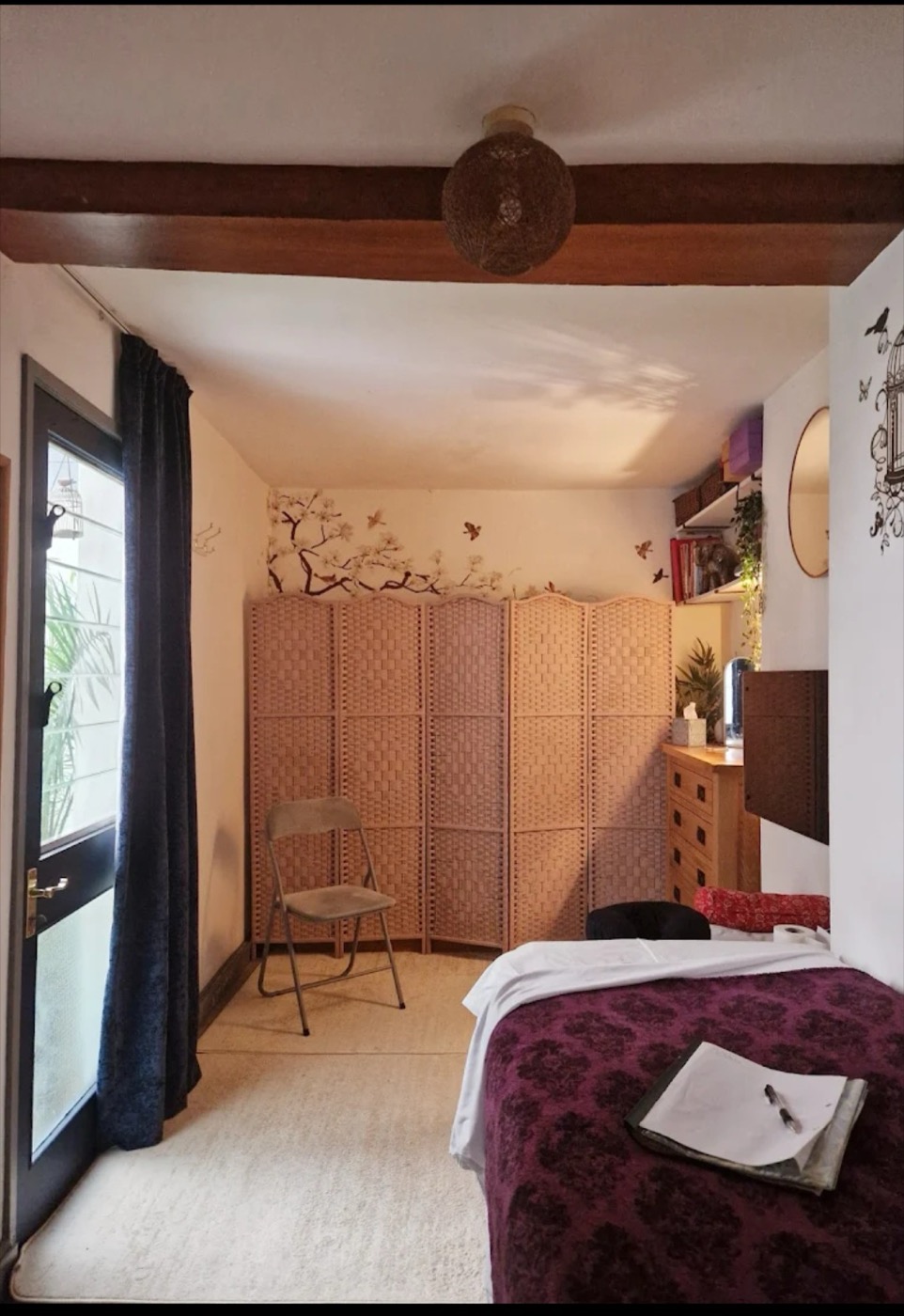 Beth's treatment room: a massage table with a deep aubergine damask throw, a wicker folding screen along the wall, and daylight through a glass door